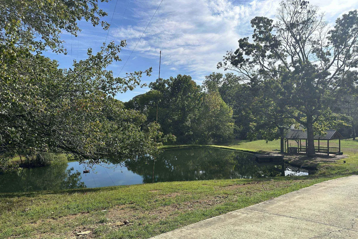 A tranquil scene of the private pond surrounded by greenery, with a large tree and a wooden gazebo visible at the water’s edge. The pond reflects the colors of sunset, and the natural setting invites fishing, picnicking, and wildlife spotting.
