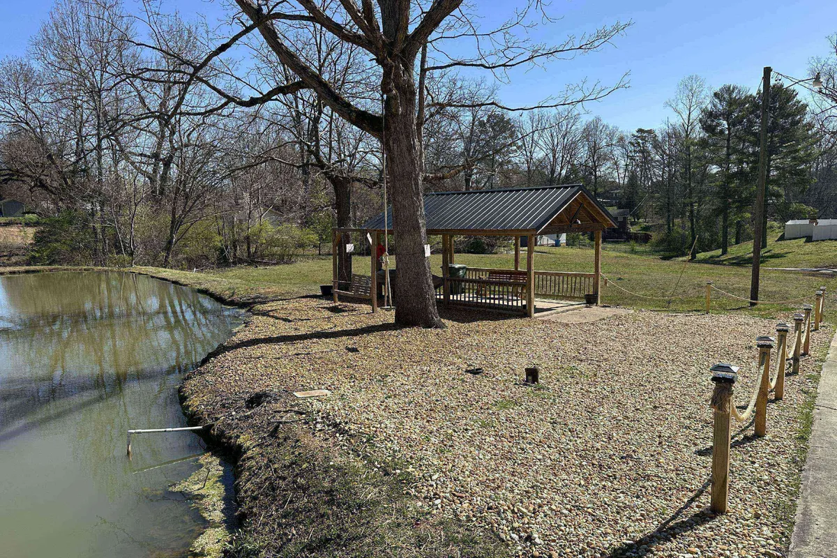 An inviting exterior scene with a gazebo overlooking a pond, accentuated by a large tree and winding gravel pathway. The area encourages peaceful nature walks, wildlife observation, and relaxing moments by the water.