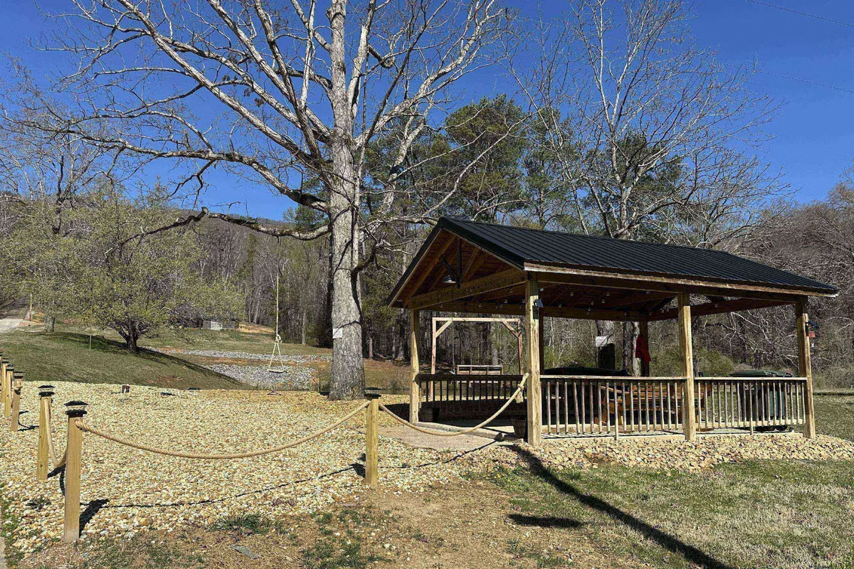 A tranquil exterior scene featuring a charming pavilion set among large trees, a gravel path, and a rope fence. The pavilion overlooks the natural landscape, providing a cozy spot for enjoying the outdoors while being surrounded by nature.