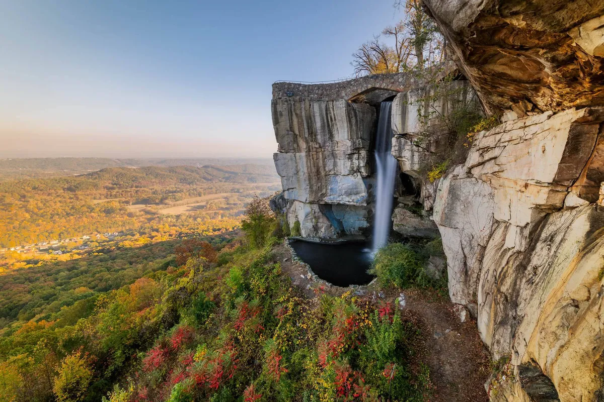 A striking exterior view featuring a dramatic waterfall cascading over a cliff edge, with expansive valley views and autumn foliage in the background. The scene captures the essence of natural exploration and scenic beauty ideal for hikers and adventurers.