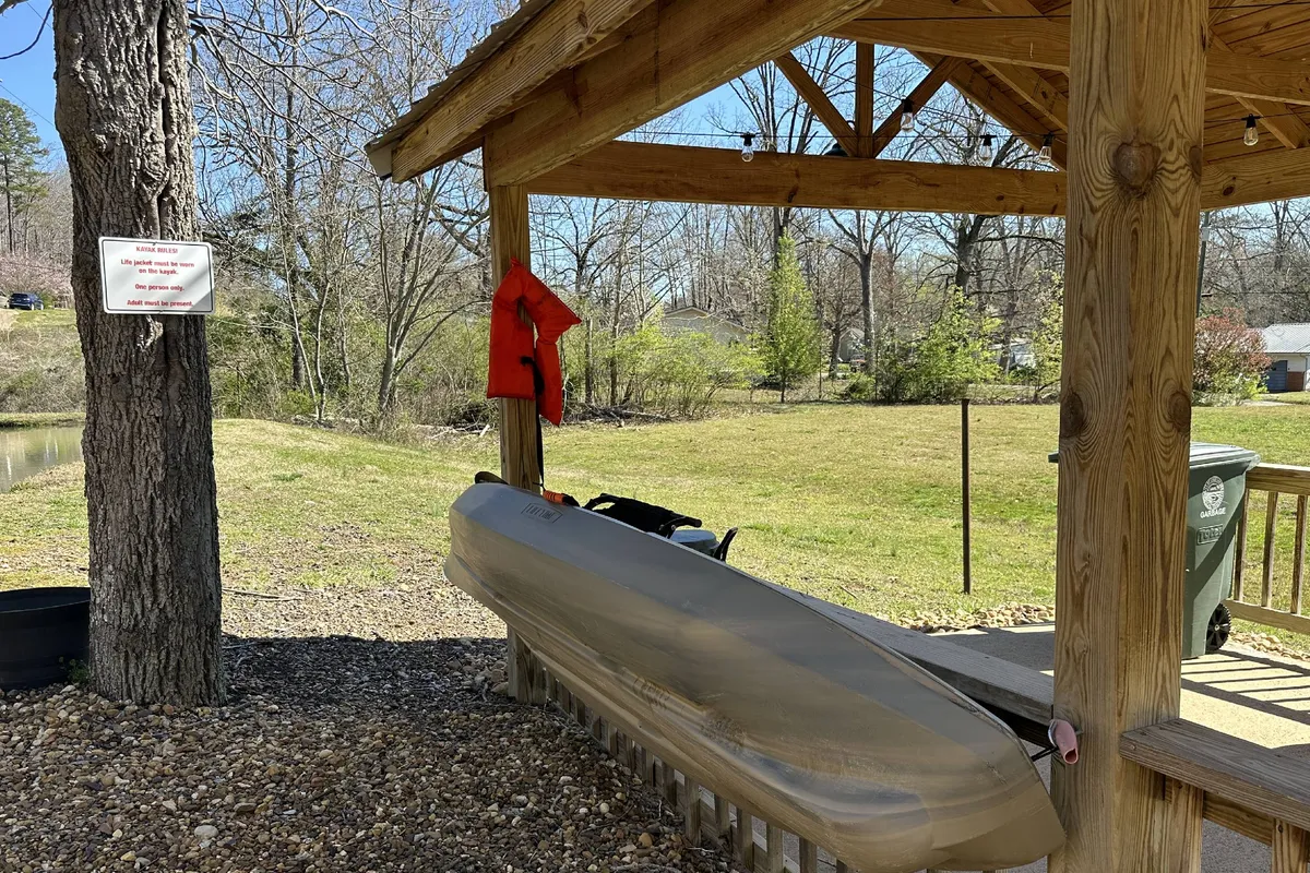 A tranquil exterior shot featuring a wooden gazebo nestled beside a calm waterway, with a kayak set out and life jackets visible. The scene captures a wide, grassy area ideal for both relaxation and outdoor play, framed by trees and open space. The sunset light adds a warm, inviting glow.