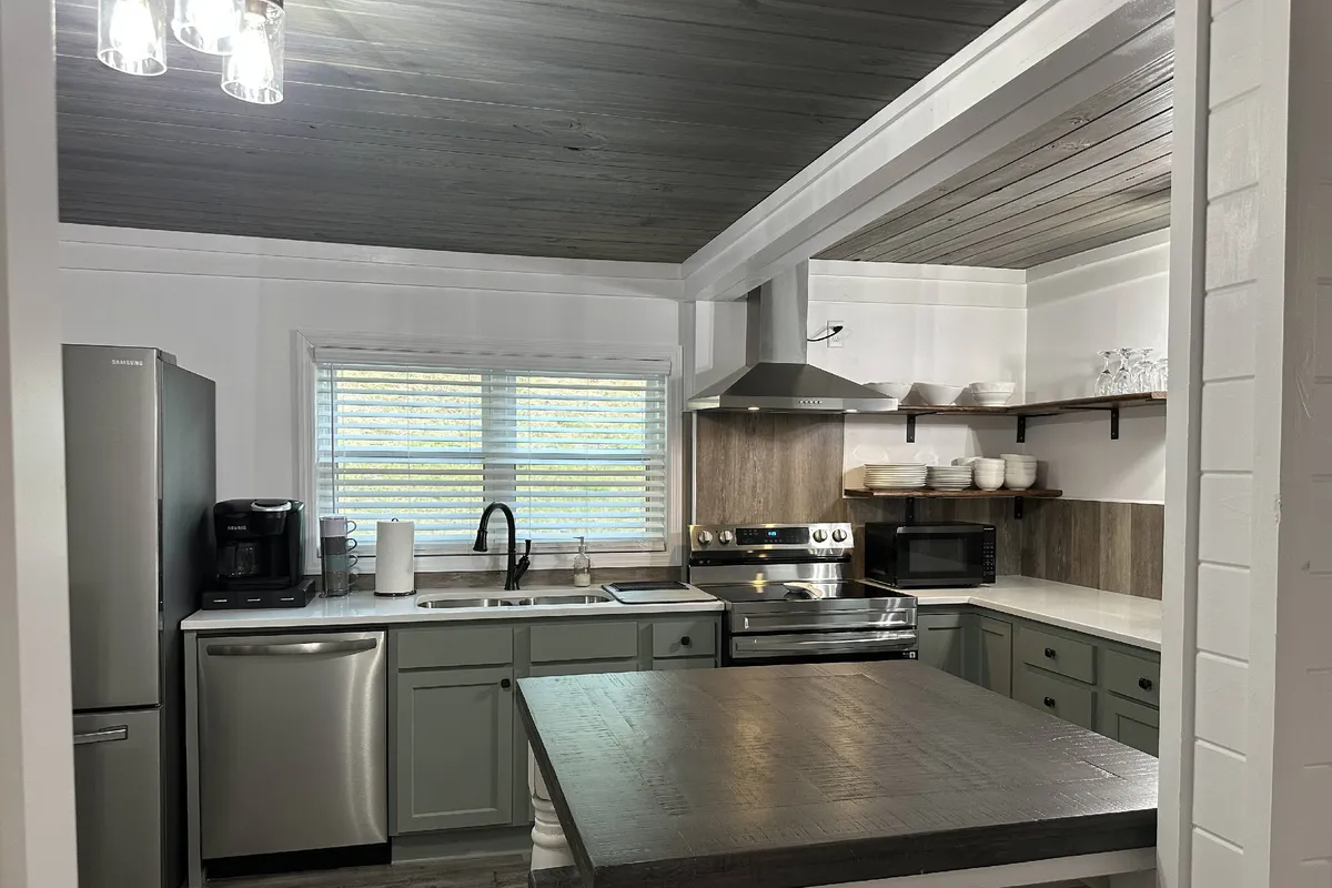 A warm and welcoming kitchen scene with a central island, stainless steel appliances, and a sunlit window. The setting suggests a space perfect for socializing while preparing meals, with ample room for more than one cook and for friends or family to gather nearby.