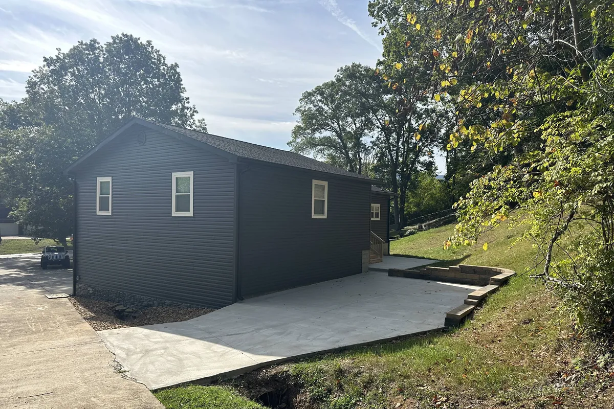 A modern home with a landscaped yard and a concrete patio set amidst verdant trees. The patio is ideal for outdoor relaxation or family gatherings, while the expansive green lawn is perfect for pets and children to play. The scene radiates early morning sunlight, enhancing the inviting atmosphere.