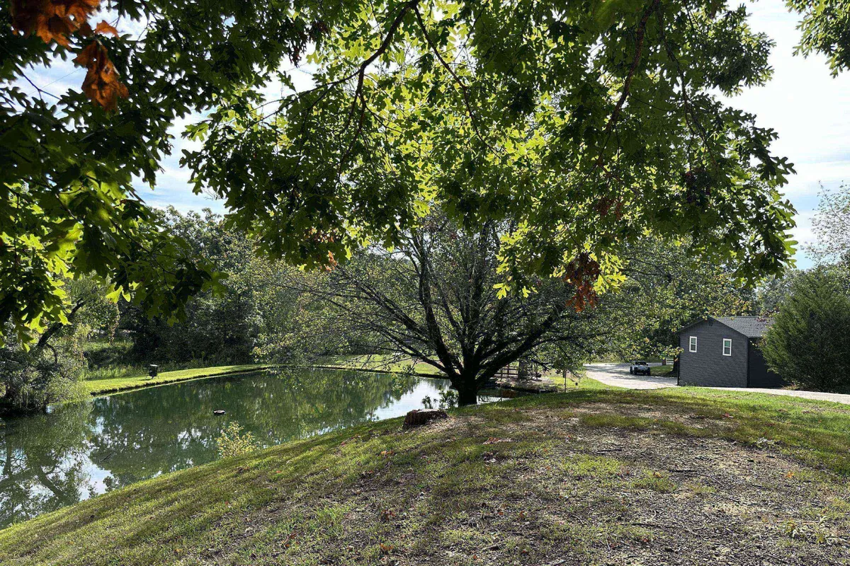 A peaceful exterior shot showing a large tree, tranquil pond, and a gray house on the bank, set amidst lush grass. The setting evokes a sense of relaxation after a day outdoors, with opportunities for walking, fishing, and watching sunsets.