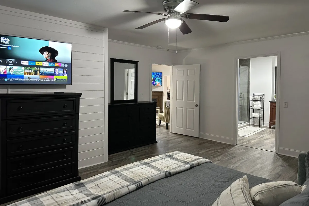 A comfortable bedroom with plaid bedding on the bed, a black dresser, a wall-mounted TV, and a mirror. The mood is cozy and the space is designed for relaxation and family time.