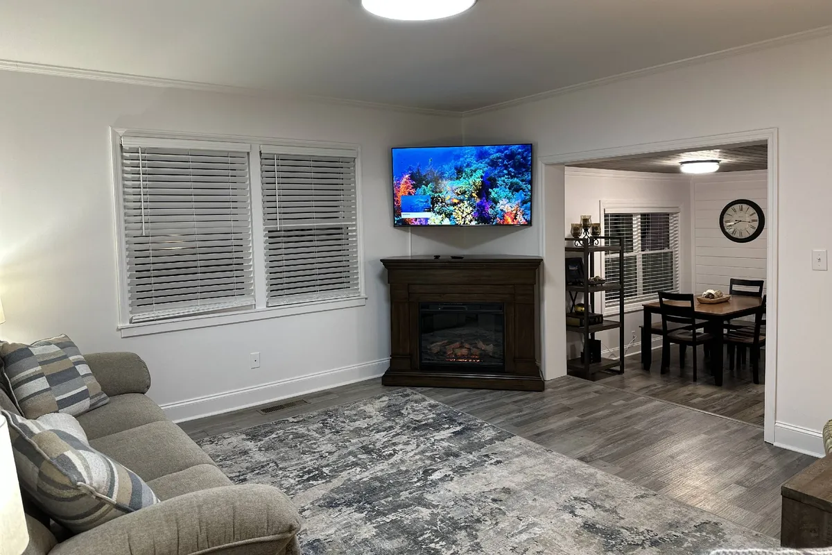 A spacious living area featuring a comfortable sofa adorned with decorative pillows, a large television displaying an underwater scene, and a cozy fireplace. Through a wide doorway, the open dining area with a large table is clearly visible, bathed in natural light from the windows.