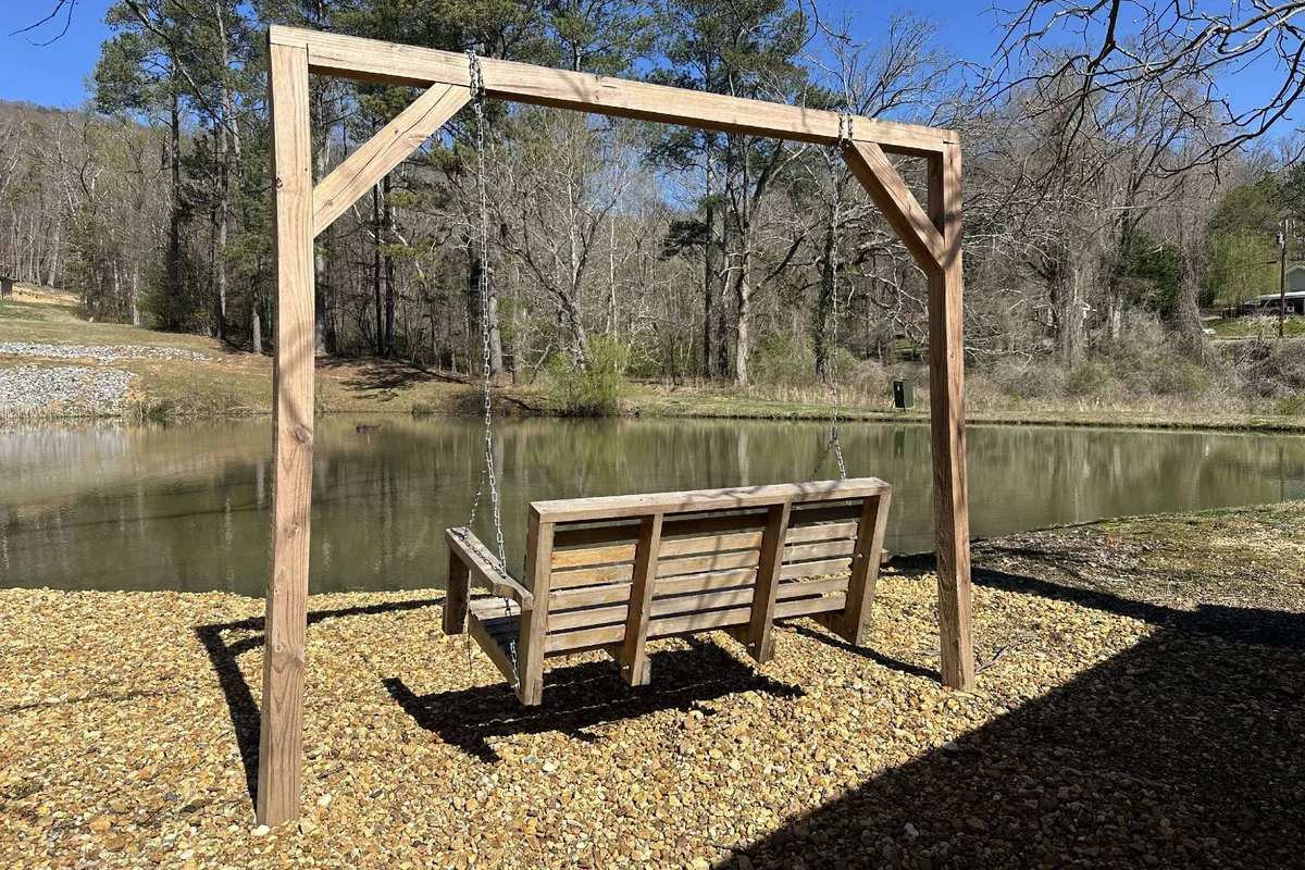 A wooden swing hangs beside a calm pond, surrounded by tall trees and lush greenery. The setting is serene, ideal for relaxing or sharing quiet moments with friends and family. Sunset colors reflect on the pond, and the natural space feels welcoming and private.